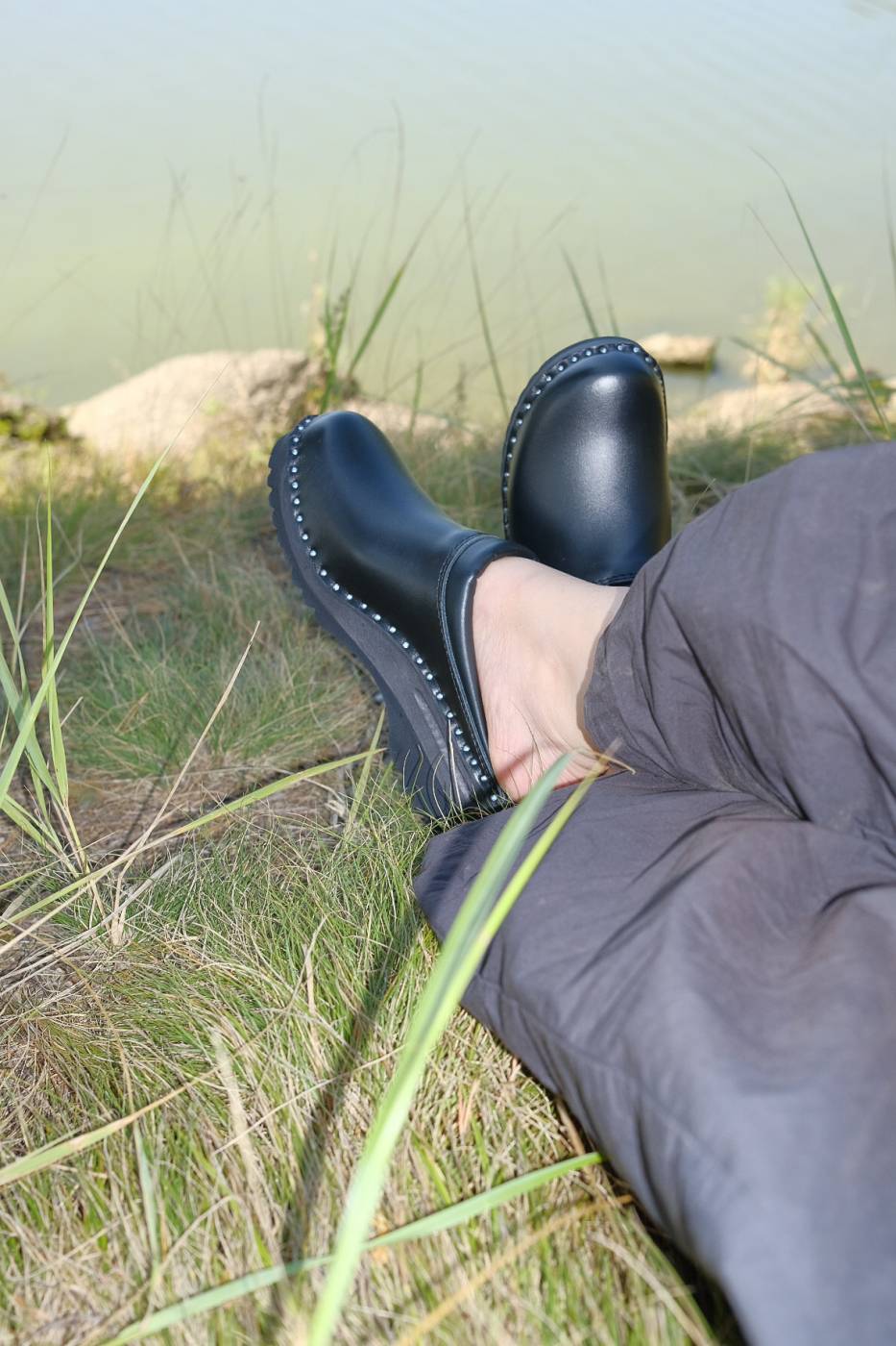 Foto von Füßen, die schwarze Holzclog Schuhe mit Oberteil aus Leder tragen. Das Lederteil ist mit silbernen Nieten an der schwarzen Holzsohle befestigt. Die Person, die die Clogs trägt, sitzt im Gras an einem Gewässer