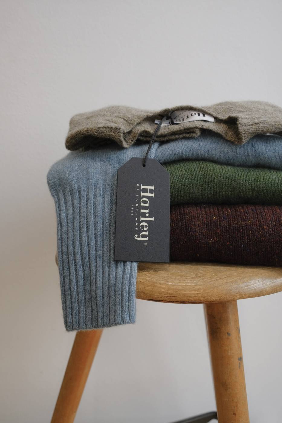 ein Hocker aus Holz mit einem Stapel von drei Stück gefalteten Wollpullovern vom schottischen Strickhersteller Harley of Scotland in den Farben Beige, Hellblau, Grasgrün und Bordeaux Rot