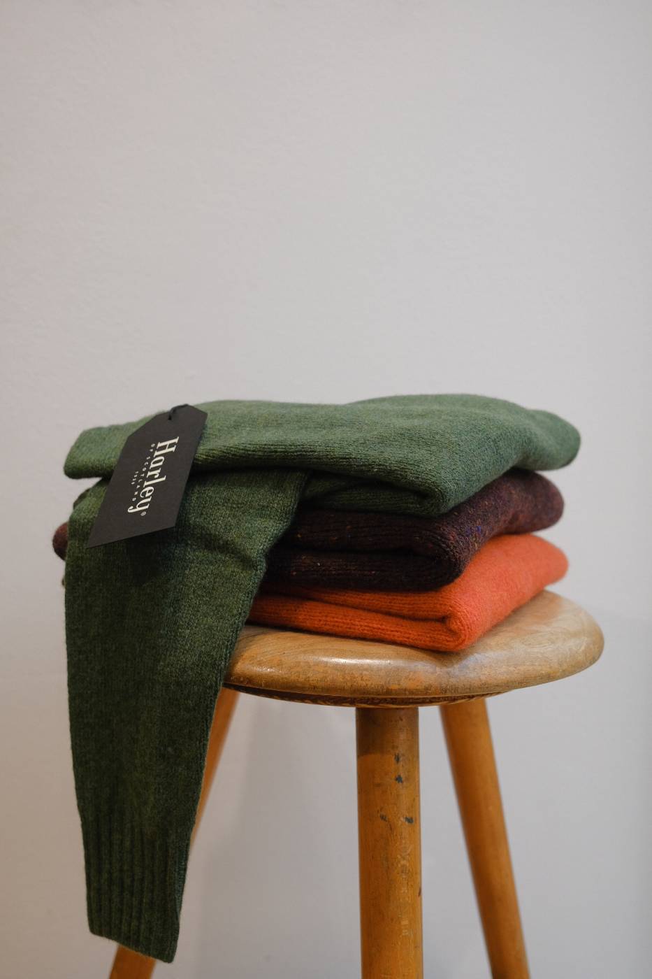 ein Hocker aus Holz mit einem Stapel von drei Stück gefalteten Wollpullovern vom schottischen Strickhersteller Harley of Scotland in den Farben Grasgrün, Bordeaux Rot und Orange